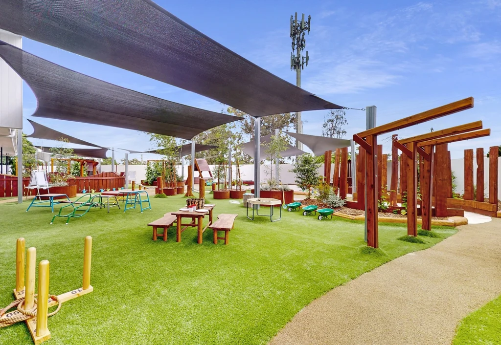 A vibrant playground with artificial turf, wooden structures, and benches under shade sails. A play area with various equipment, including a seesaw and climbing frames, is visible.