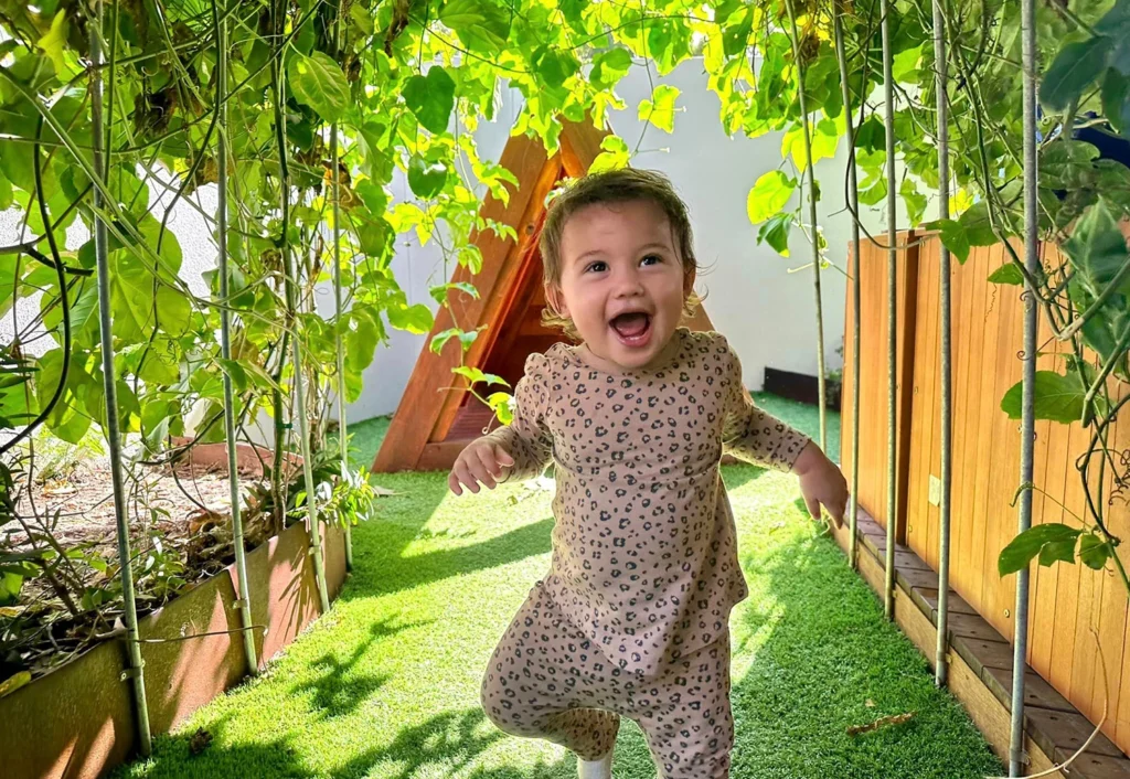 A young child wearing a leopard print outfit smiles joyfully while walking under a leafy garden archway.