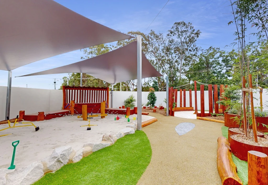 A children's outdoor play area with a sandpit, play equipment, shaded sails, and landscaped garden beds. There are wooden logs and structures arranged around the space. Trees are visible in the background.