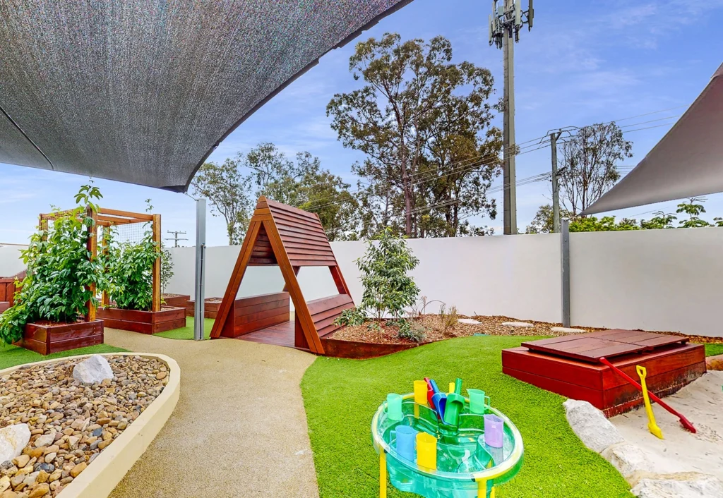 A playground features a wooden climbing structure, raised garden beds, a sandpit with toys, and a water play station, all under shade sails with trees and a blue sky in the background.