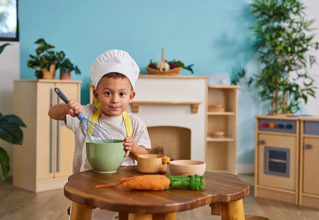 A young child wearing a chef's hat is stirring ingredients in a bowl at a table with vegetables and kitchen items in a play kitchen setting.