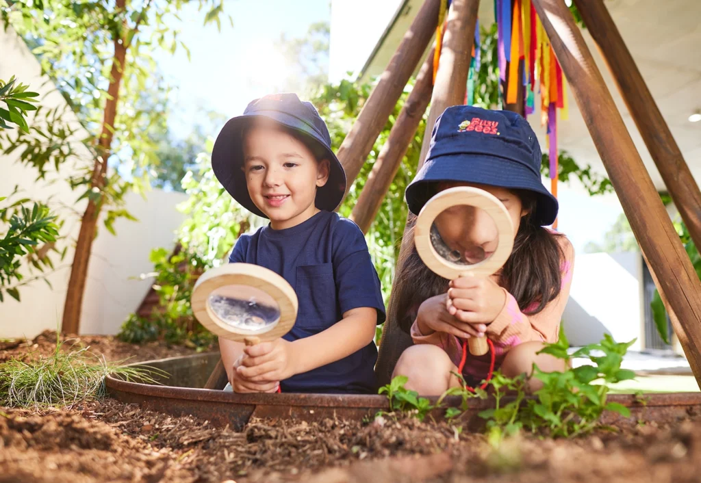 Two children wearing hats and holding magnifying glasses kneel in a garden next to a wooden teepee structure. Bright sunlight can be seen through the trees behind them.