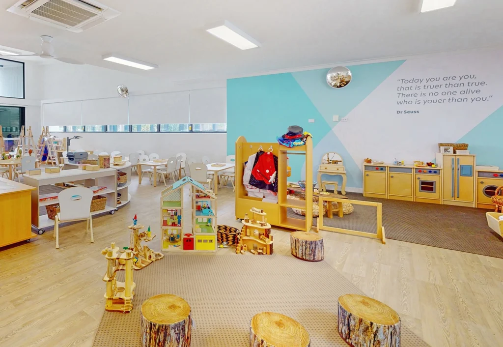 A brightly lit classroom with wooden play structures, stools made from tree stumps, a play kitchen, and child-sized tables and chairs. A wall has a quote by Dr. Seuss.