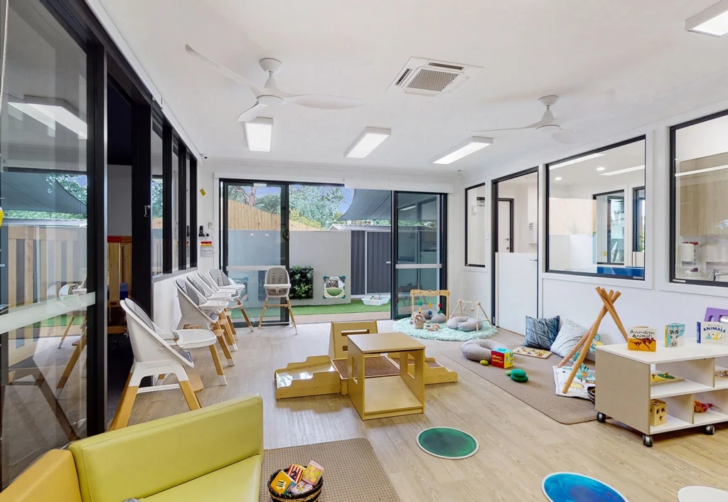 A bright daycare room with toys, books, chairs, and play structures. Large windows and glass doors provide natural light and a view of the outdoor play area. Ceiling fans and modern lighting are visible.