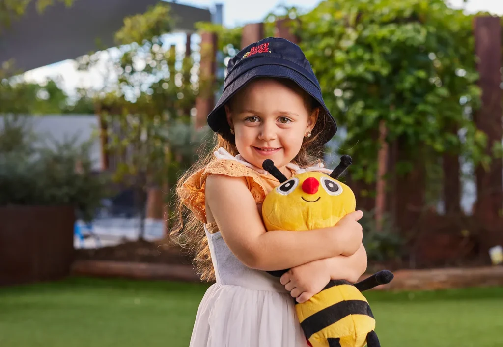 A young girl wearing a dark hat and a light dress smiles while hugging a plush bee toy outside in a garden.