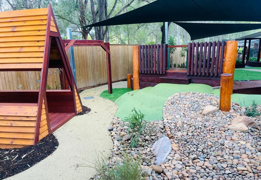 Outdoor playground featuring wooden climbing structures, green artificial turf hills, a dry rock bed, and shaded areas under large canopies, surrounded by a wooden fence and trees in the background.