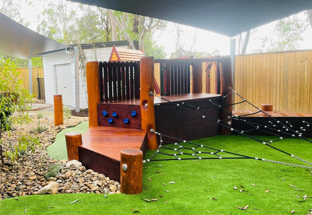 A children’s outdoor play area featuring a wooden climbing structure, rope nets, and artificial turf, located under a shaded canopy with a wooden fence in the background.