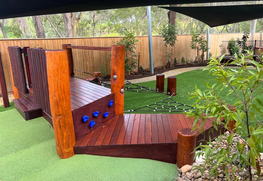 Outdoor playground area featuring a wooden climbing structure with blue handholds and a rope bridge, surrounded by artificial grass and shaded by a canopy.