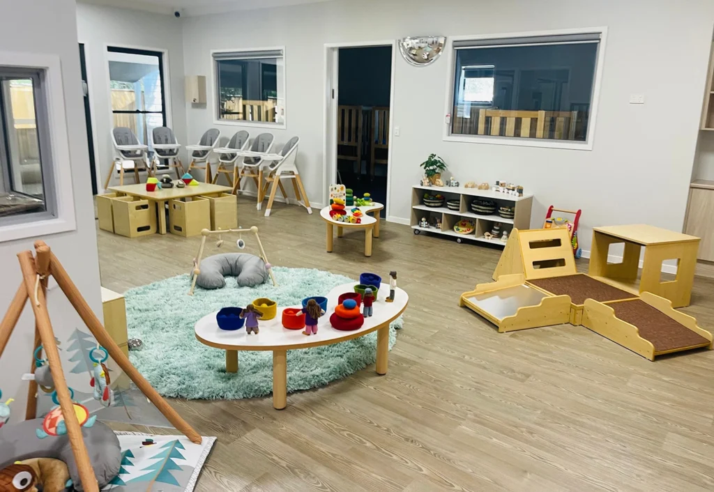 A brightly lit, organized daycare room with various toys, play structures, and small furniture. There are several high chairs against the wall and various educational materials on shelves.