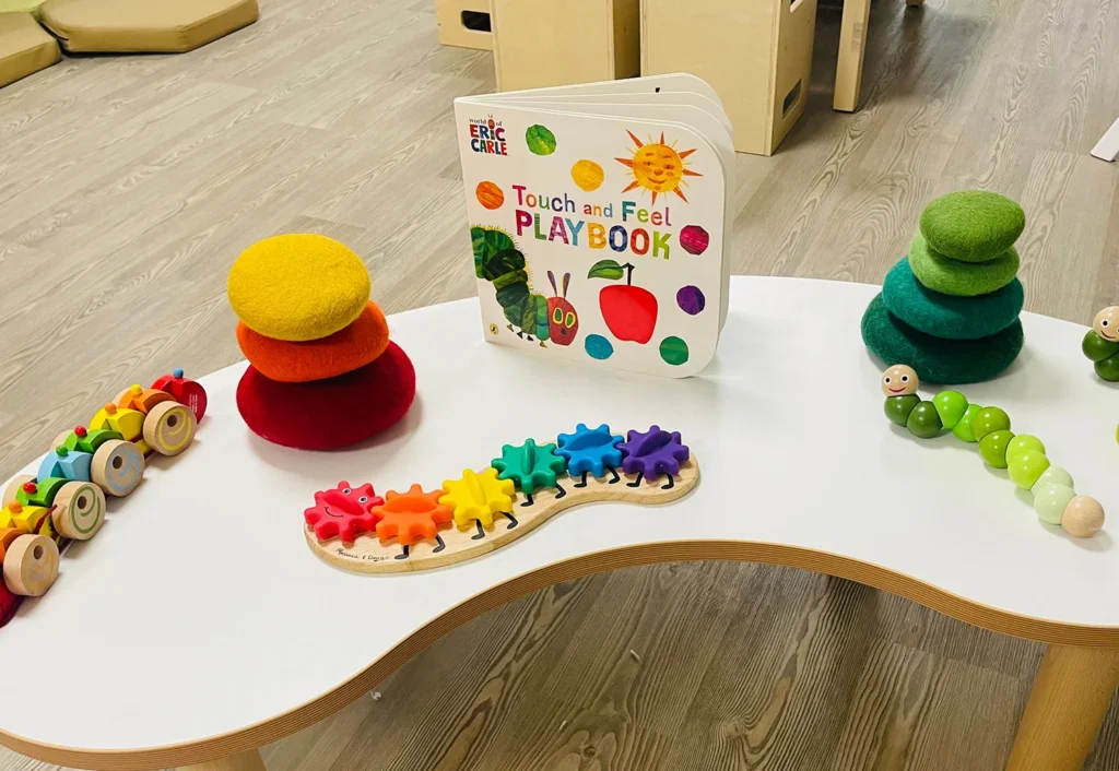 A white table featuring colorful children's toys including a caterpillar toy, a gear puzzle, stackable felt discs, and a "Touch and Feel Playbook" by Eric Carle.