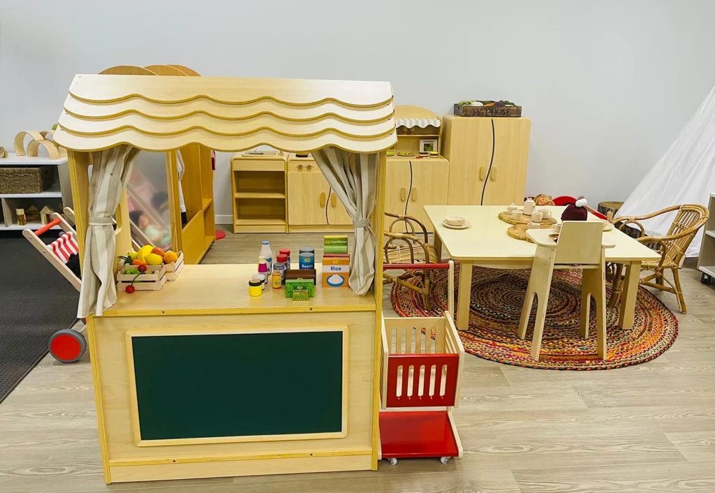 A children's play area with a toy market stand, shelves of toy food items, a cart, and a table with chairs in a brightly lit room.