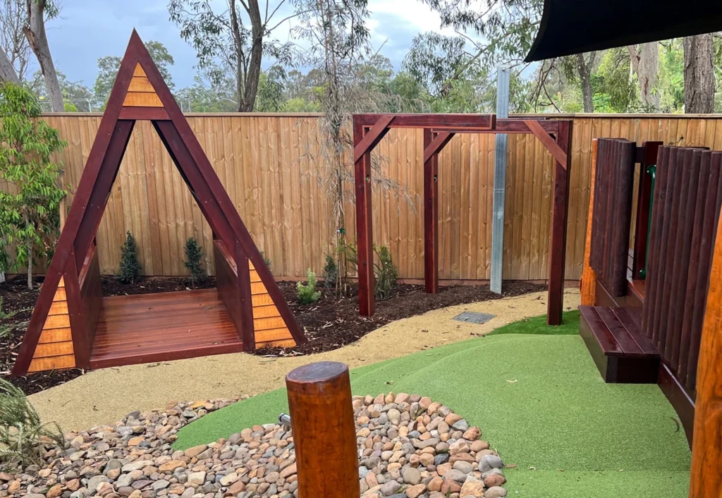 A wooden children's play area features a triangular frame, a small climbing structure, and a shaded bench on artificial turf, surrounded by a wooden fence and trees in the background.