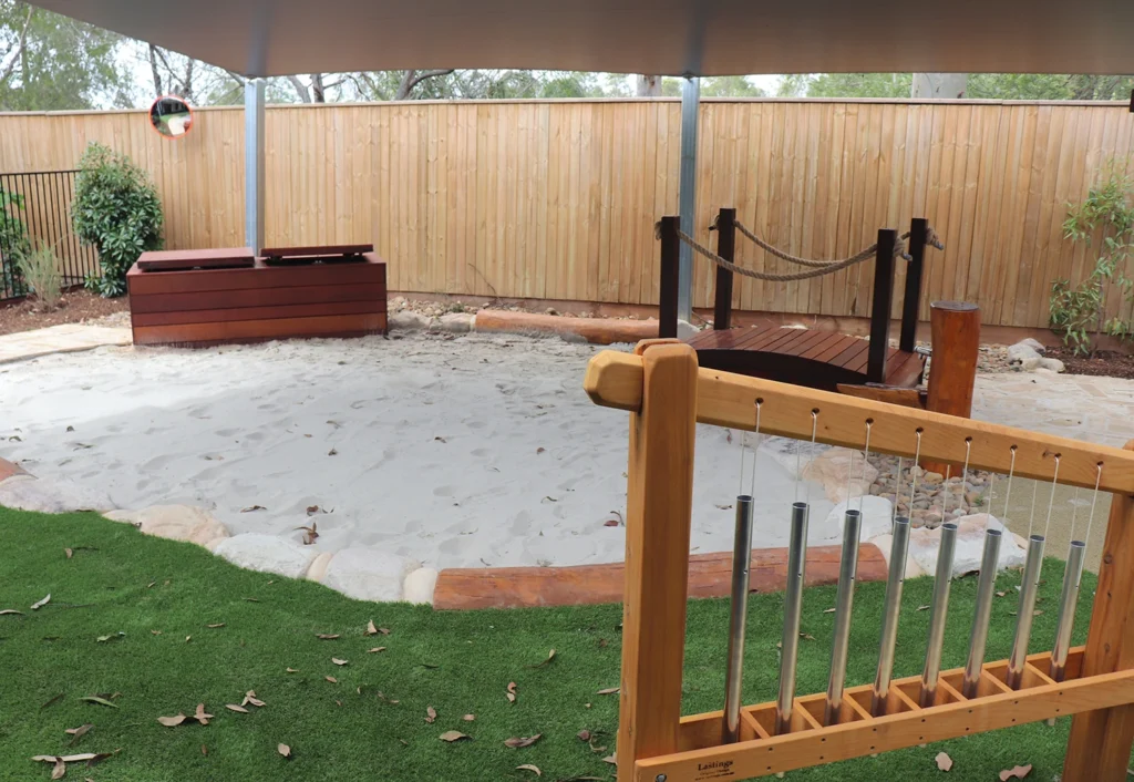 Outdoor play area featuring a sandbox, wooden bridge, climbing wall, and metal chimes, surrounded by a wooden fence and shaded by a roof.