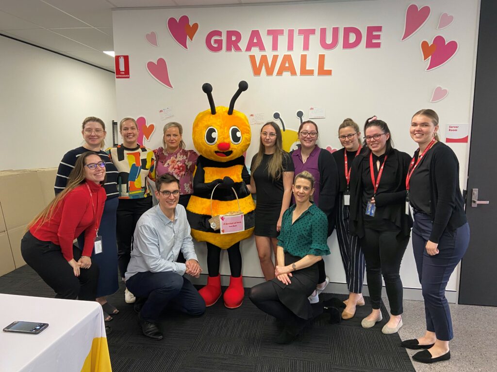 A group of people posing with a person in a bee costume in front of a "Gratitude Wall" decorated with heart shapes.