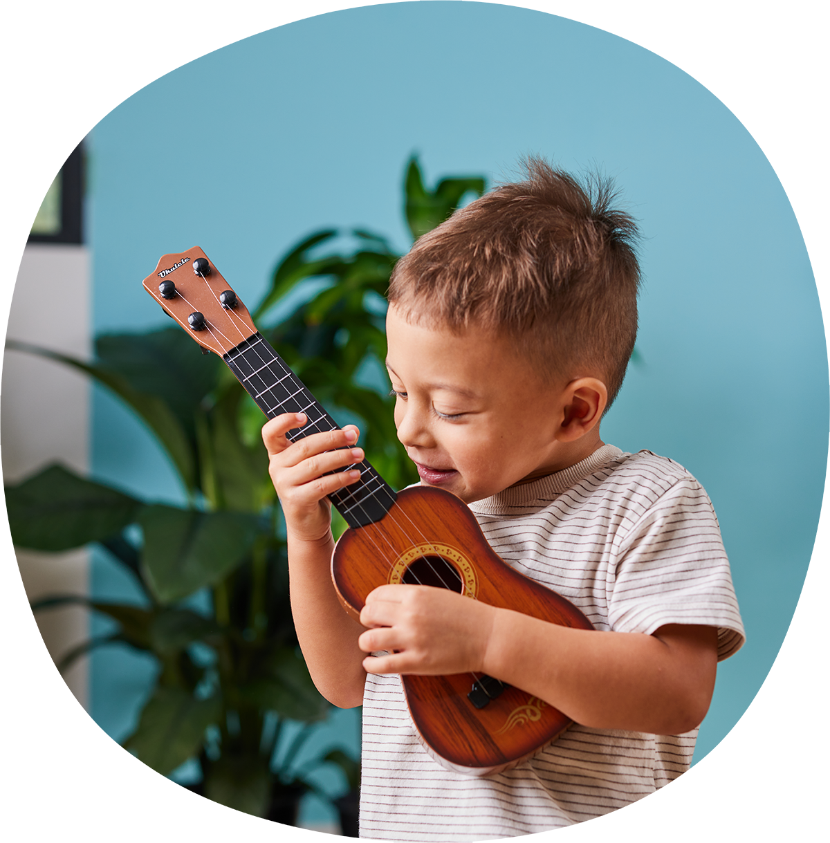 A young boy with short hair is playing a small wooden ukulele. He is wearing a striped t-shirt and standing in front of a blue wall with green plants in the background.