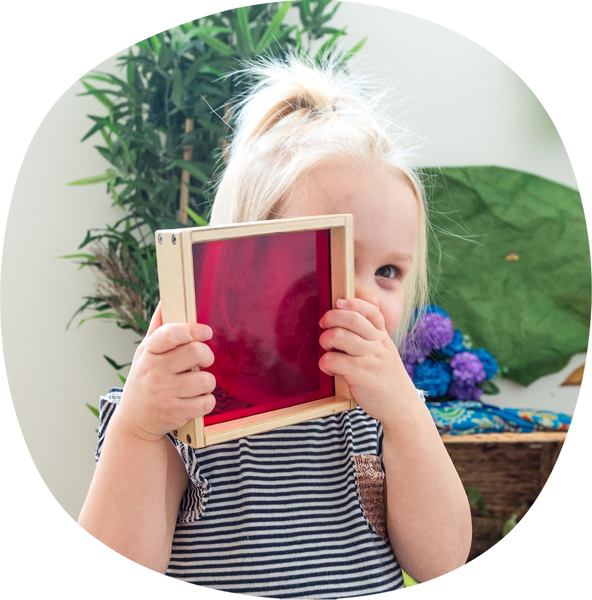 A child with blonde hair holds a wooden frame with a red translucent screen up to their face. Green foliage and colorful objects are in the background.