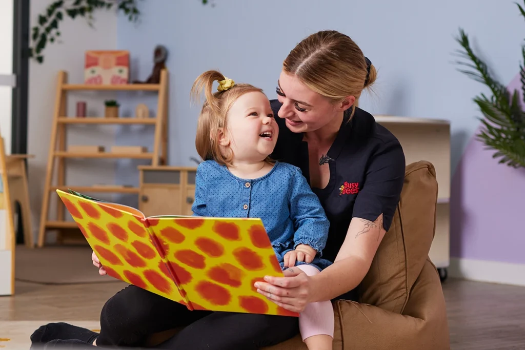 A woman smiles and reads a large colorful book to a toddler, who also smiles, in a room with shelves and plants.