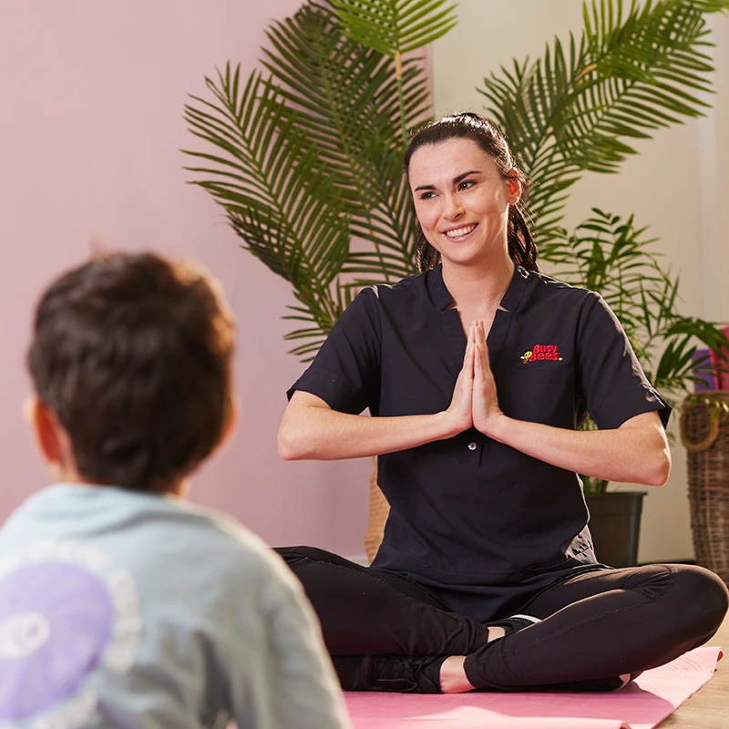 A woman sits cross-legged on a mat with hands in a prayer position, smiling at a child seated opposite her. There are green plants in the background.