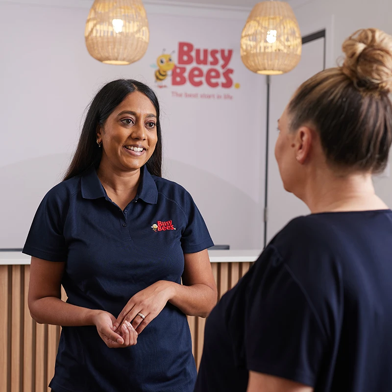 Two women are talking in an office with a "Busy Bees" sign on the wall. One woman is wearing a navy polo shirt with the "Busy Bees" logo.