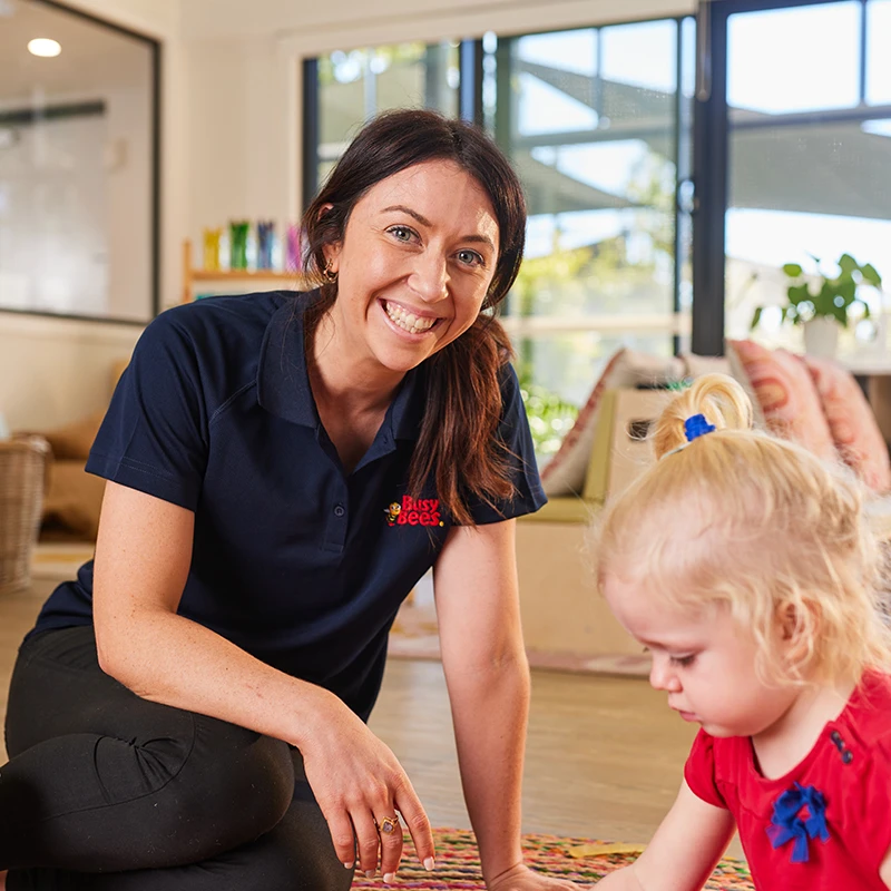 A woman in a navy blue polo shirt with a company logo smiles while kneeling next to a young child playing, in a bright room with large windows.