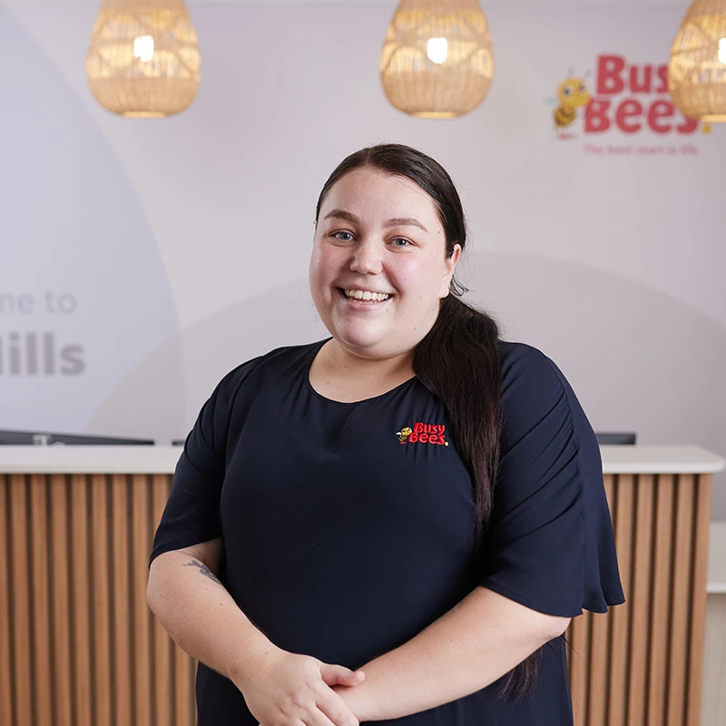 A person with long dark hair standing and smiling in front of a reception desk at a Busy Bees childcare center.