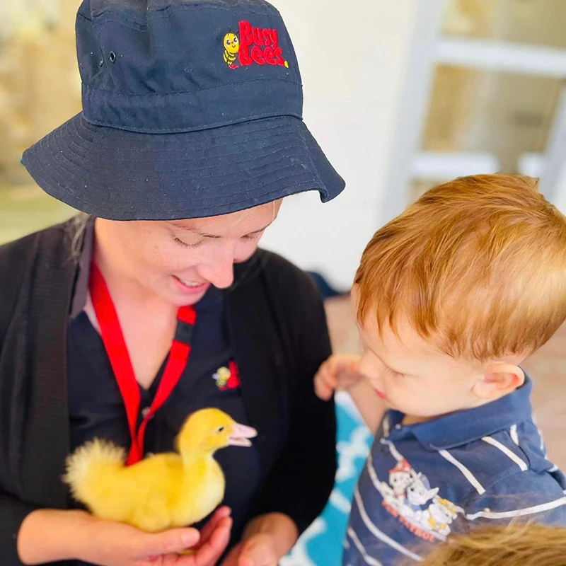 A woman wearing a black hat with a "Busy Bees" logo holds a yellow duckling, while a young child looks at the duckling.