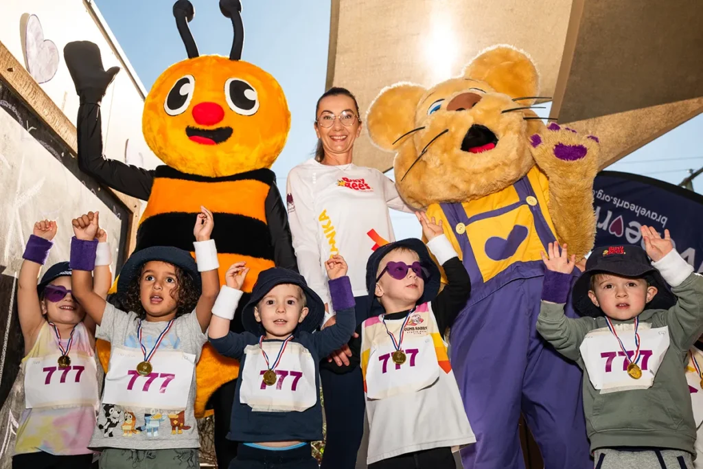 Busy Bees Early Learning's mascot "Buzz the bee" with Bravehearts' mascot "Ditto the lion cub" along with young children with their hands up celebrating
