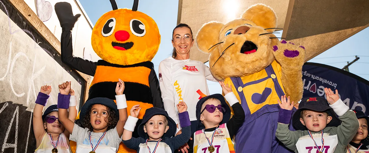 Busy Bees Early Learning's mascot "Buzz the bee" with Bravehearts' mascot "Ditto the lion cub" along with young children with their hands up celebrating