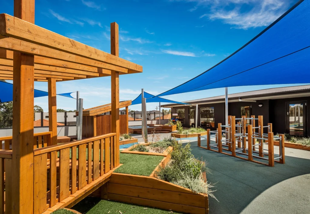 Outdoor playground with wooden structures and blue shade sails under a clear sky. The area features grassy and paved surfaces with various play equipment.