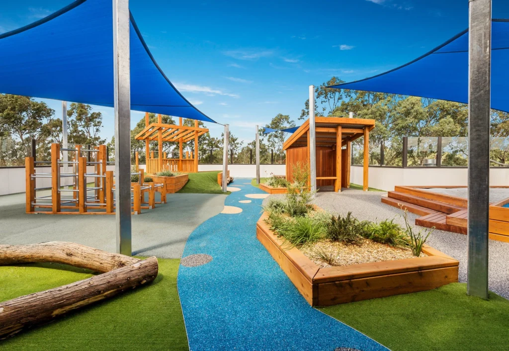 Outdoor playground with various play structures, wooden shelters, and blue shade sails. The ground is covered with a mix of green turf and blue rubberized paths, with nature-inspired elements.