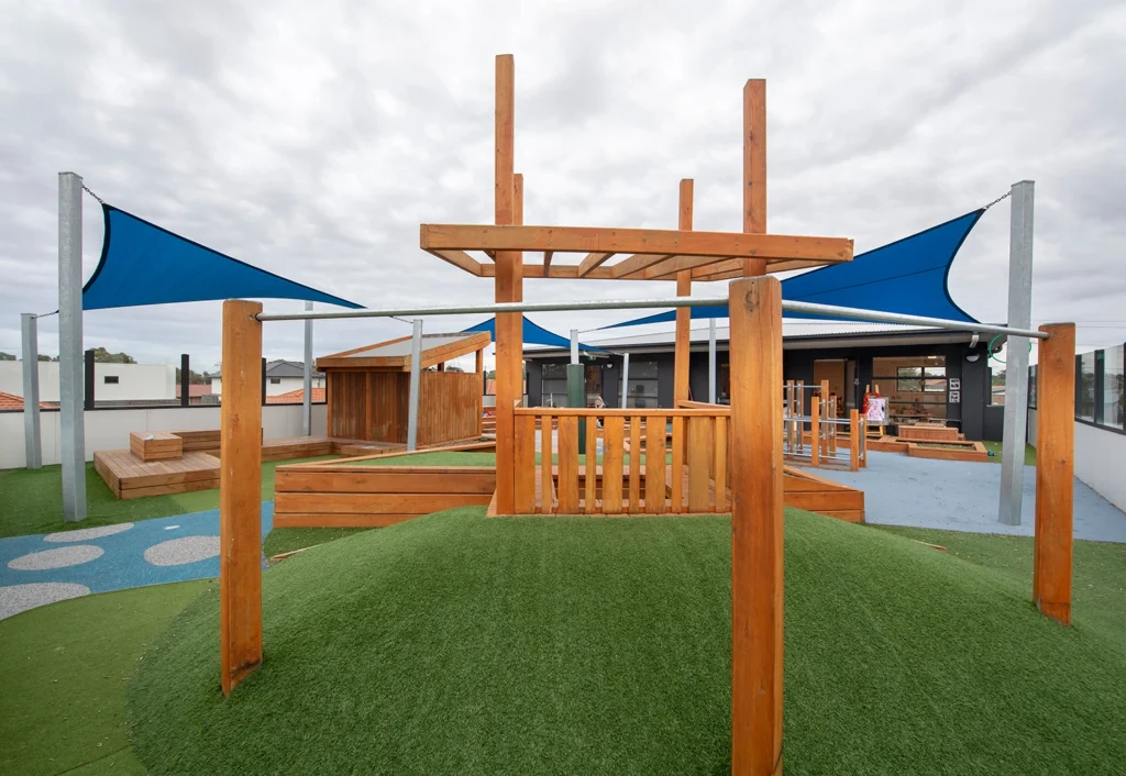 Outdoor playground with wooden climbing structures, blue shade sails, and artificial turf under a cloudy sky.