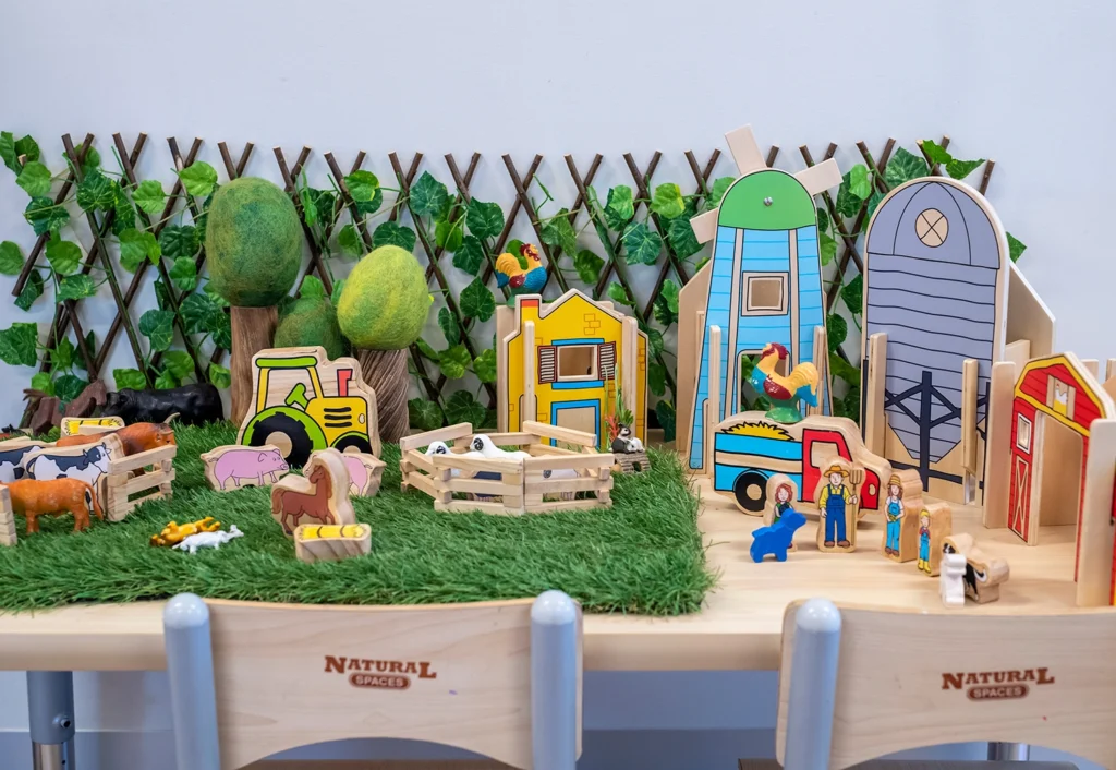 A tabletop farm playset with wooden animals, structures, and a toy tractor, set against a green backdrop with artificial grass and foliage.