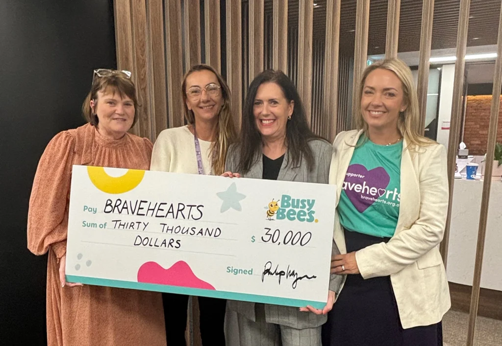 Four women stand holding a large $30,000 check from Busy Bees to Bravehearts, smiling at the camera.