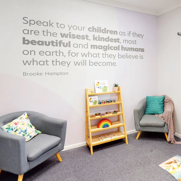 A cozy reading nook with two grey armchairs, a wooden shelf with children's books, and a motivational quote on the wall that reads, "Speak to your children as if they are the wisest, kindest, most beautiful and magical humans on earth, for what they believe is what they will become".