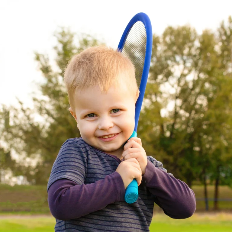 A young child with blond hair, smiling while holding a blue tennis racket over their shoulder, enjoys a day in the greenery, reflecting the joy and innocence of kindergarten.