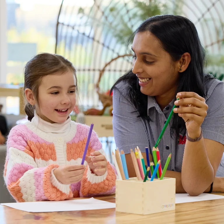 A child and an adult sit at a table in the lively childcare, smiling and holding colored pencils. A box of pencils is on the table in front of them, highlighting the joy of creative activities in this nurturing day care environment.