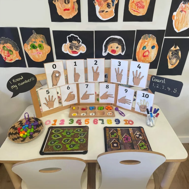 An educational setup ideal for a preschool includes hand-drawn number posters, finger counting cards, sorting toys, and activity boards displayed on a table, with children's artwork adorning the wall behind.