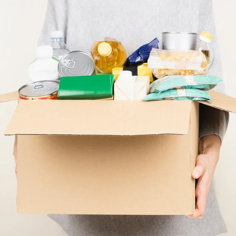 A person holding a cardboard box filled with various food items, including cans, bottles and packages.