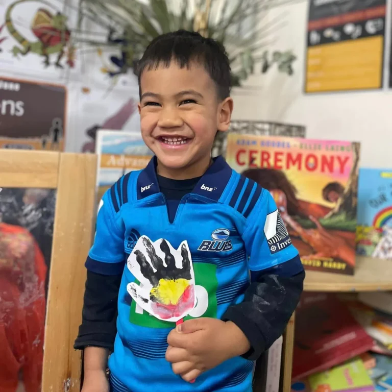 A young child in a blue rugby jersey smiles while holding a craft with a painted handprint. There are book covers visible in the background, capturing the joy of early learning in kindergarten.