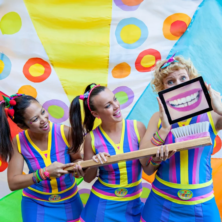 Three women in colorful outfits hold a large toothbrush and a tablet displaying a smiling mouth against a vibrant backdrop of circles and stripes, perfect for promoting early learning in kindergarten or day care settings.