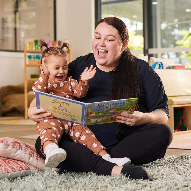 A woman and a child sit on the floor looking at a book together. The child, excited with hands raised, eagerly listens as the woman points to the book and smiles. They are in a bright, cozy daycare room.