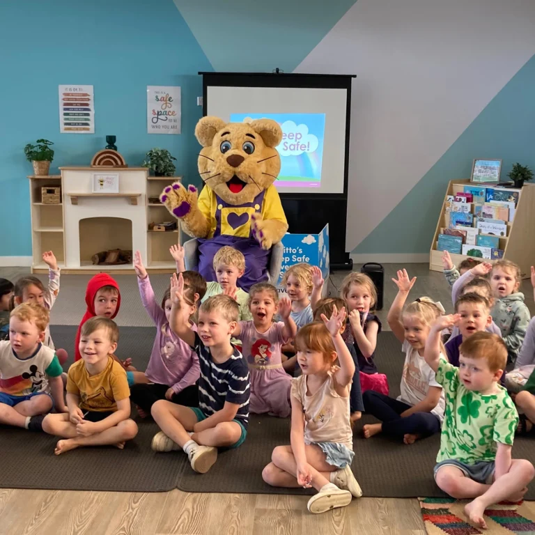 A group of kindergarten children sit on the classroom floor, smiling and raising their hands, with a person in a lion cub costume sitting behind them in front of a projection screen that says "Keep Safe!".