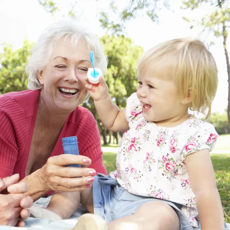 An elderly woman and a young child are sitting on grass. The woman smiles while the child holds a bubble wand, appearing to have fun outdoors in a nurturing childcare setting.