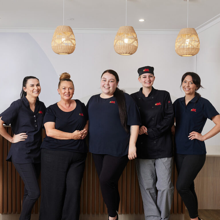 Five women in matching navy blue uniforms stand in a row inside a modern room with pendant lights.