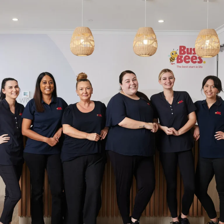 Six women in matching navy blue uniforms stand in a row inside a modern room with pendant lights and a sign that reads "Busy Bees Day Care.