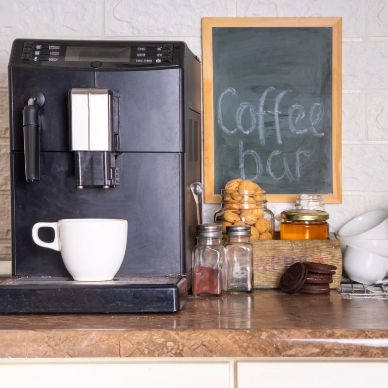 A coffee machine is placed on a kitchen counter next to a white mug, jars of cookies, spices, and a chalkboard sign that reads "Coffee bar." Nearby, colorful preschool art projects add a touch of early learning charm to the cozy setting.