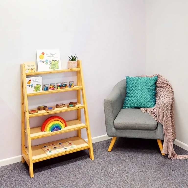 Image of a cozy reading nook perfect for early learning, featuring a wooden shelf holding children's books, jars, and toys. Adjacent is a gray armchair with a teal pillow and a pink throw. Ideal for any kindergarten or childcare setting.