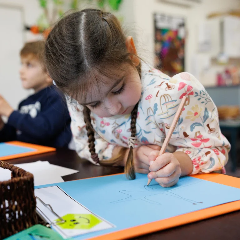 A young girl with braided hair draws on blue paper at a day care classroom table, with another child visible in the background.