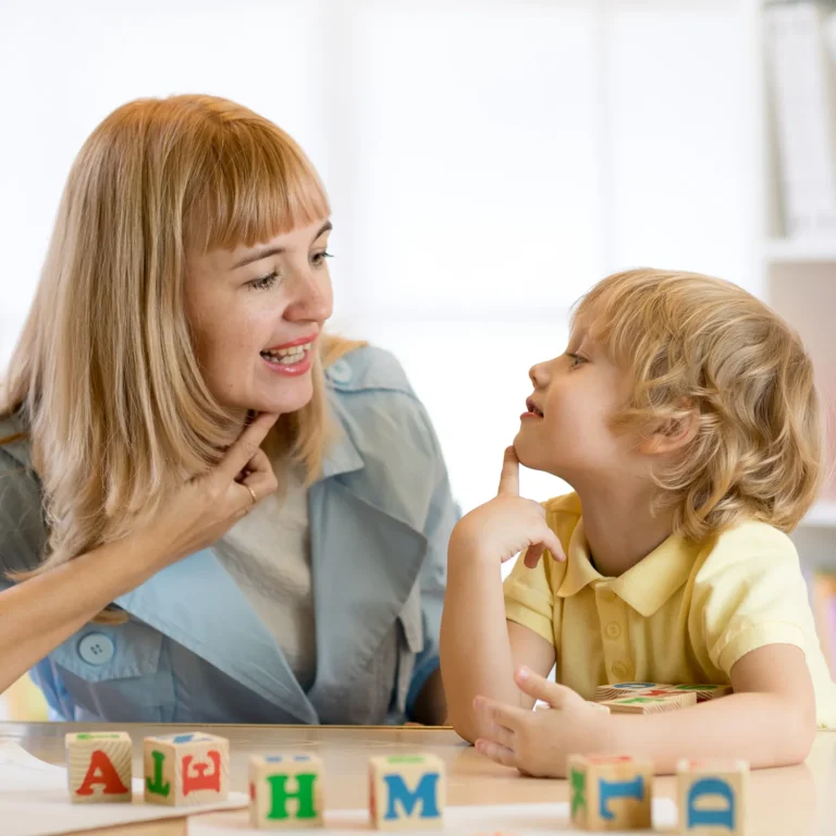 A woman and a young boy engage in early learning at a table with alphabet blocks. The boy, immersed in their kindergarten-style activity, points to his chin while looking at her.