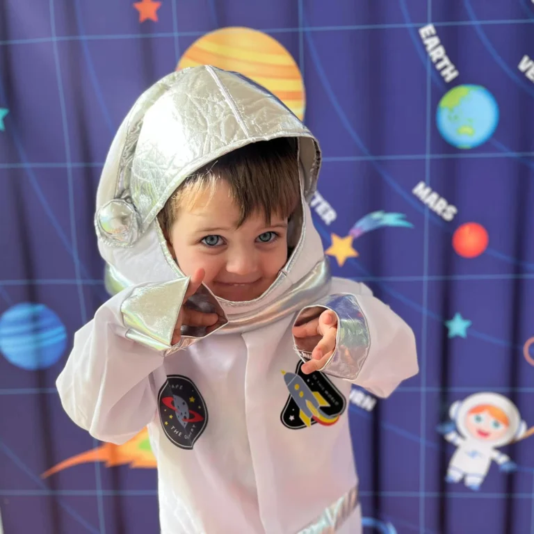 A preschool child dressed in a silver astronaut costume points forward against a space-themed backdrop with planets and stars.